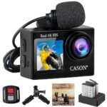 A black CASON CS6 action camera featuring a front-facing color display and a large lens. The camera is shown with its included accessories: a clip-on external microphone with a foam windscreen, a wrist-mounted remote control, a mini tripod mount, a user manual, and a dual-slot battery charger containing two rechargeable batteries.